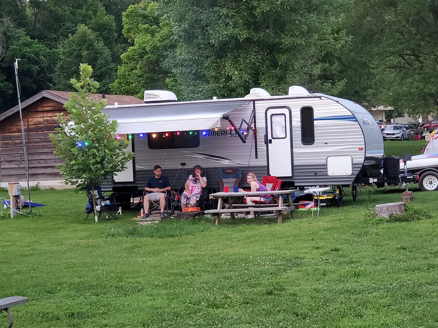 Family relaxing outside a camper trailer at a campsite, with chairs, a table, and a barbecue grill, surrounded by trees.