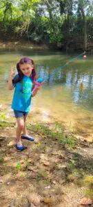 A young girl with purple hair ties poses by a creek, proudly holding up a fishing rod with a small fish attached.