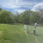 Lush green park with trees under a clear blue sky, featuring a chain-link fence enclosing a grassy area and a pathway alongside it.