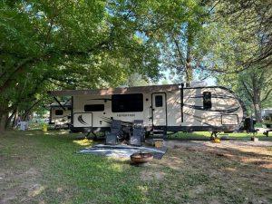 A recreational vehicle (rv) parked in a shaded campsite with trees, featuring an extended awning and a small fire pit in front.