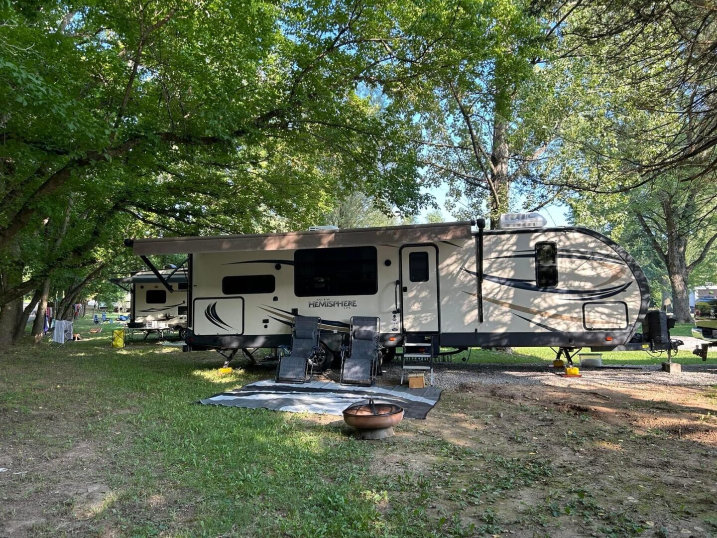A recreational vehicle (rv) parked in a shaded campsite with trees, featuring an extended awning and a small fire pit in front.