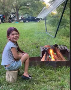 A young girl smiling while sitting on a stump next to a campfire in a park, with vehicles and trees in the background.