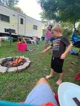 A young boy in a batman shirt stands near a campfire in a grassy area with a camper and other people in the background.
