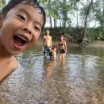 A young boy smiling at the camera, with three other children playing in a shallow river, surrounded by trees.