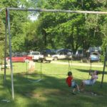 Two children on a swing set in a sunny park with cars and trailers in the background.